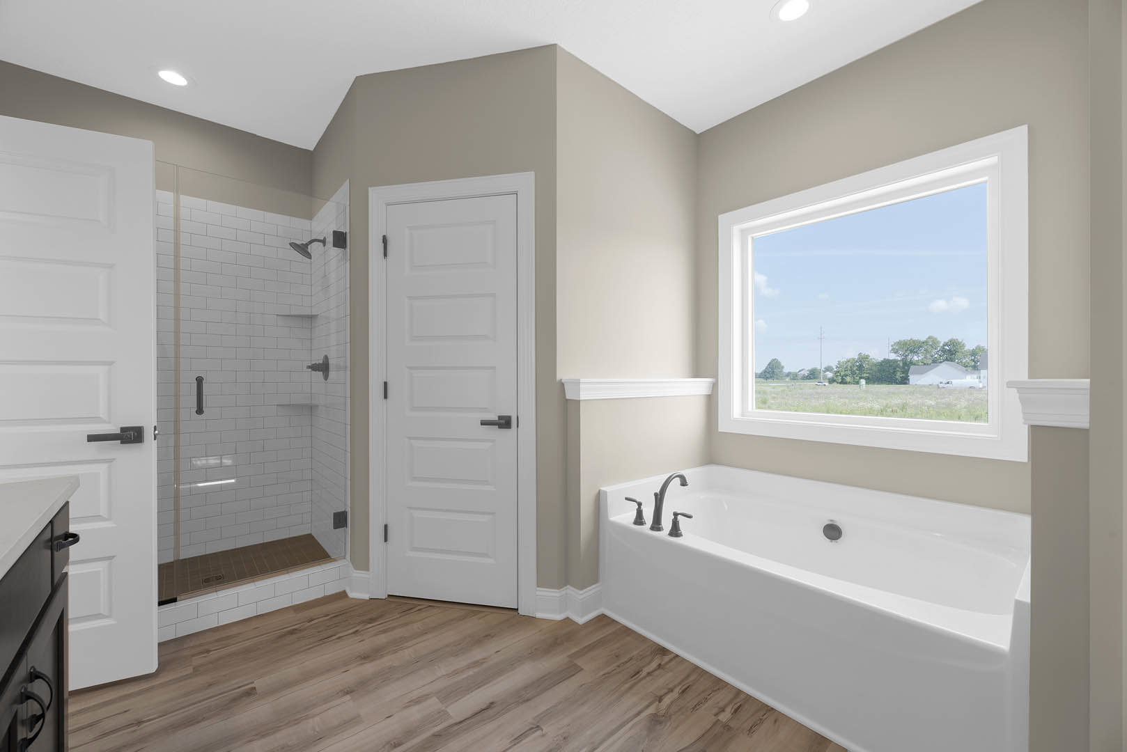 Bathroom featuring a freestanding bathtub with chrome faucet on wood flooring, glass-enclosed shower with tile walls, white door with black handle, and window overlooking a field