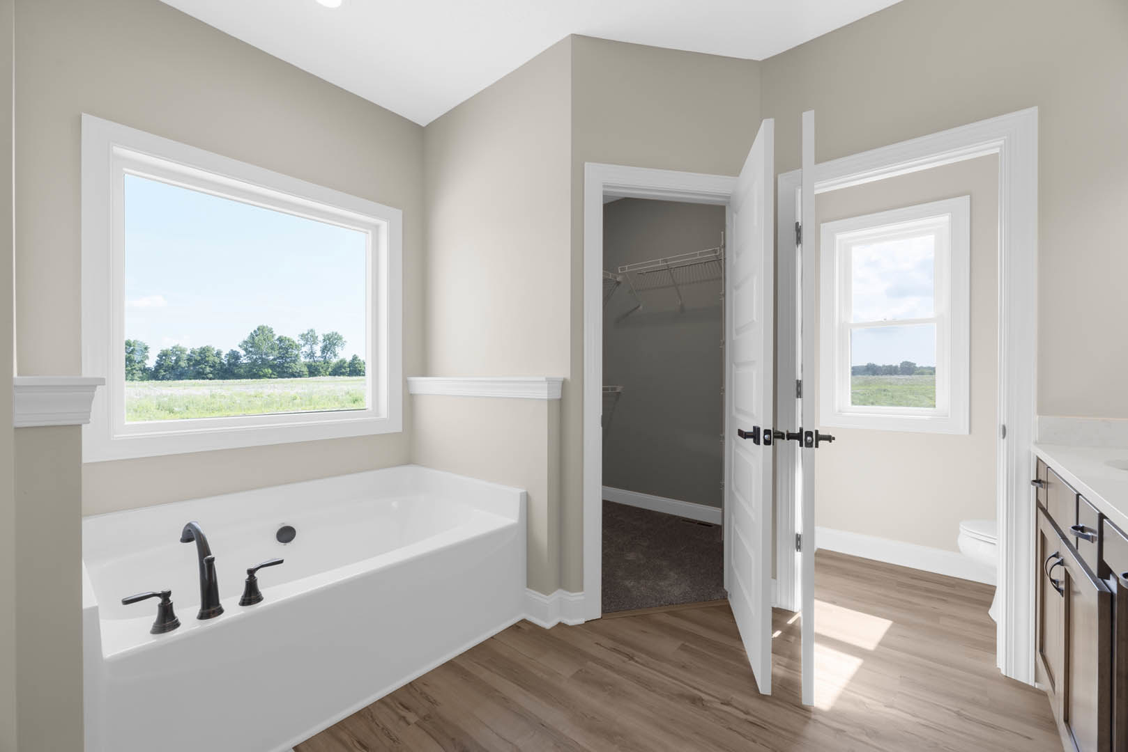 Bathroom with freestanding bathtub, chrome faucets, wood floor, open door, large window showing trees and field, white walls and ceiling