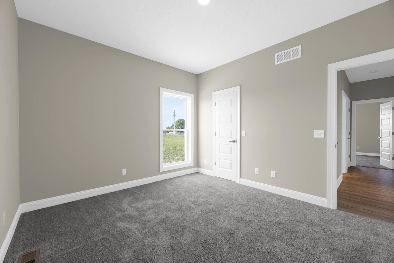 White door with silver handle beside a large window overlooking a grassy field, white plaster walls, beige carpet, and hardwood laminate flooring