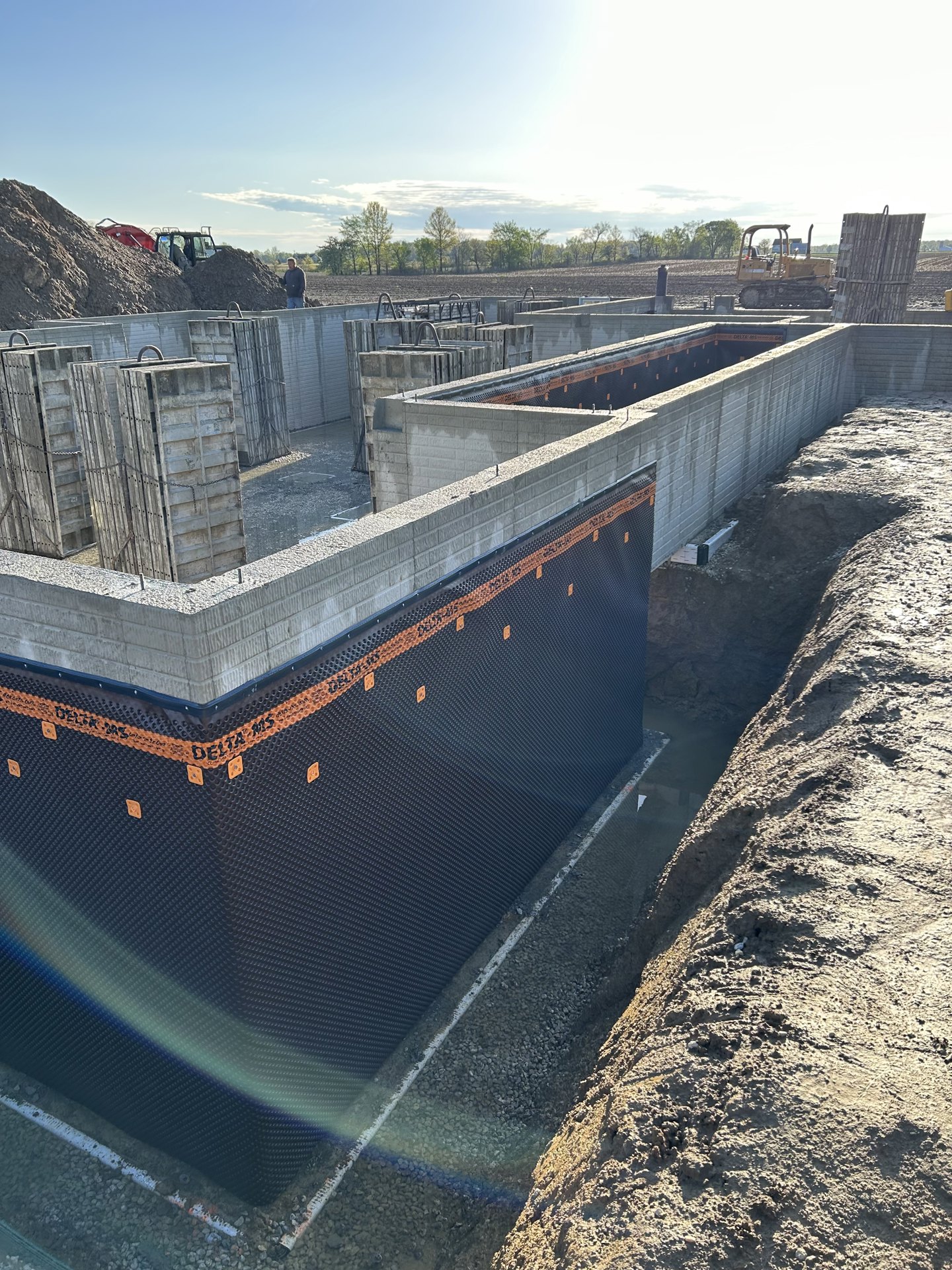 Large concrete foundation surrounded by dirt, stacked concrete blocks, and brick walls; bulldozer parked on a dirt road; trees visible in the background under an open sky.