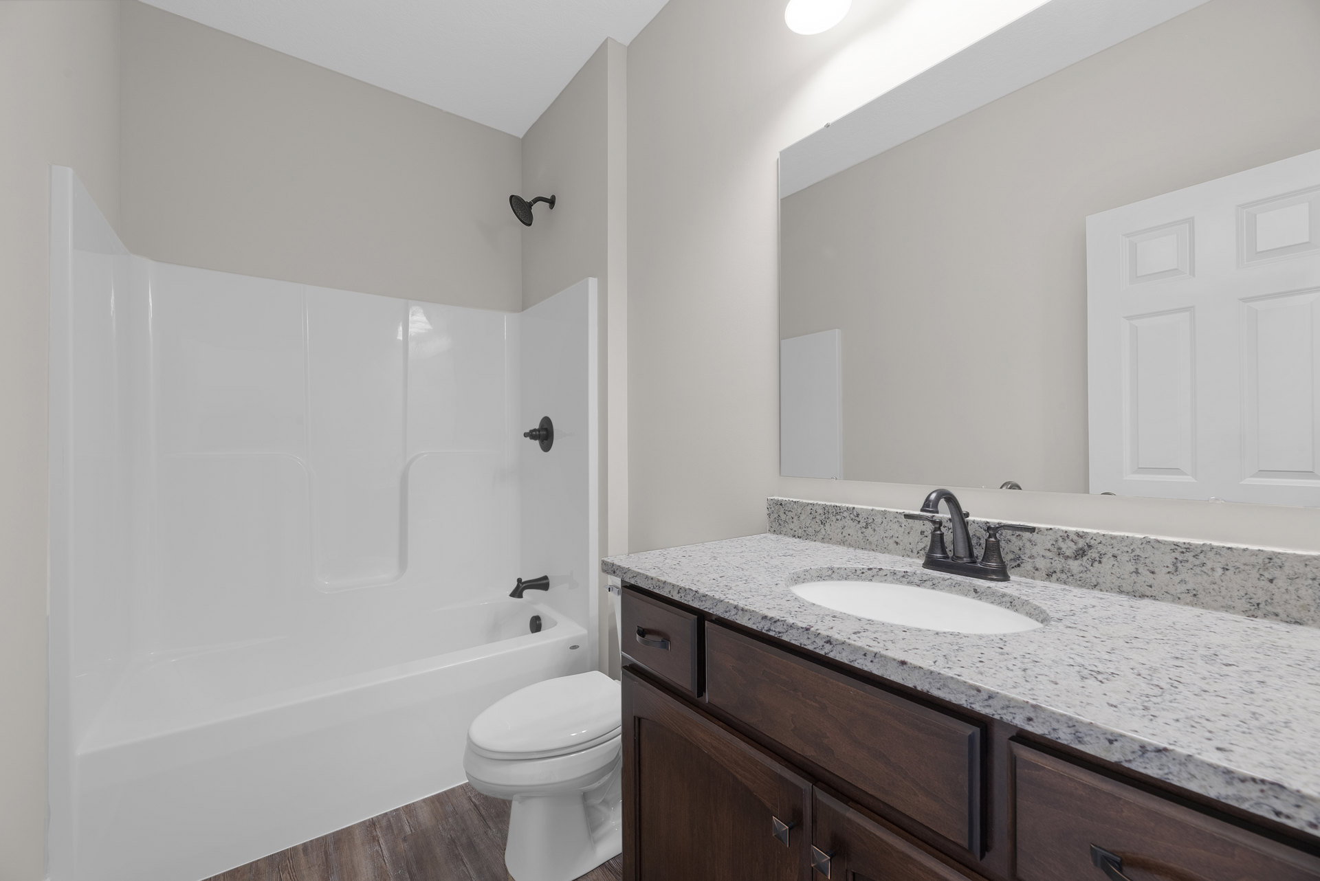 Bathroom with white bathtub, closed-lid toilet, wall-mounted shower head, light-colored tile walls, sink set in a stone countertop, and wood cabinetry.