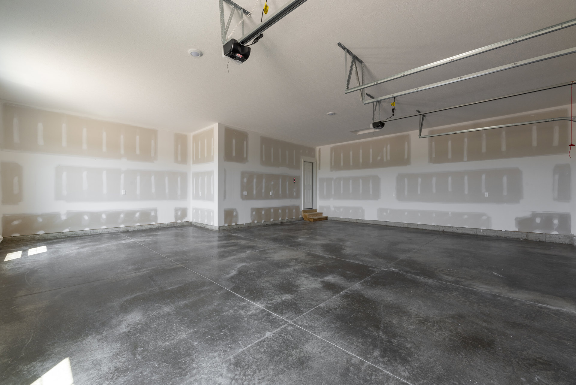 Concrete floor with white walls, ceiling-mounted projector, metal ceiling frame, and staircase; minimalist interior with grey tile flooring and visible white lines.