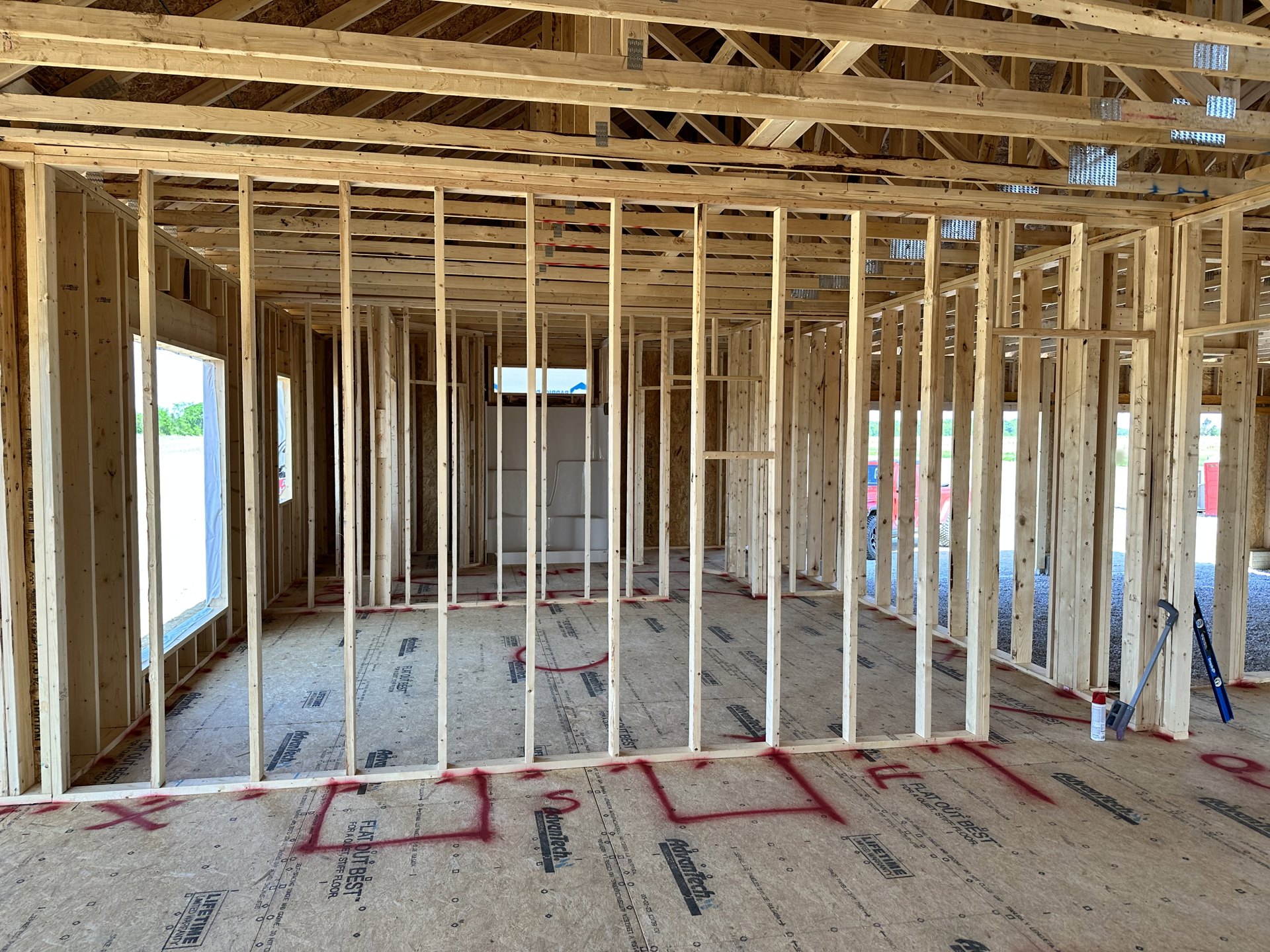 Exposed wooden beams and framing structure inside a house under construction, unfinished plywood floor with red markings, blue level tool resting on the ground