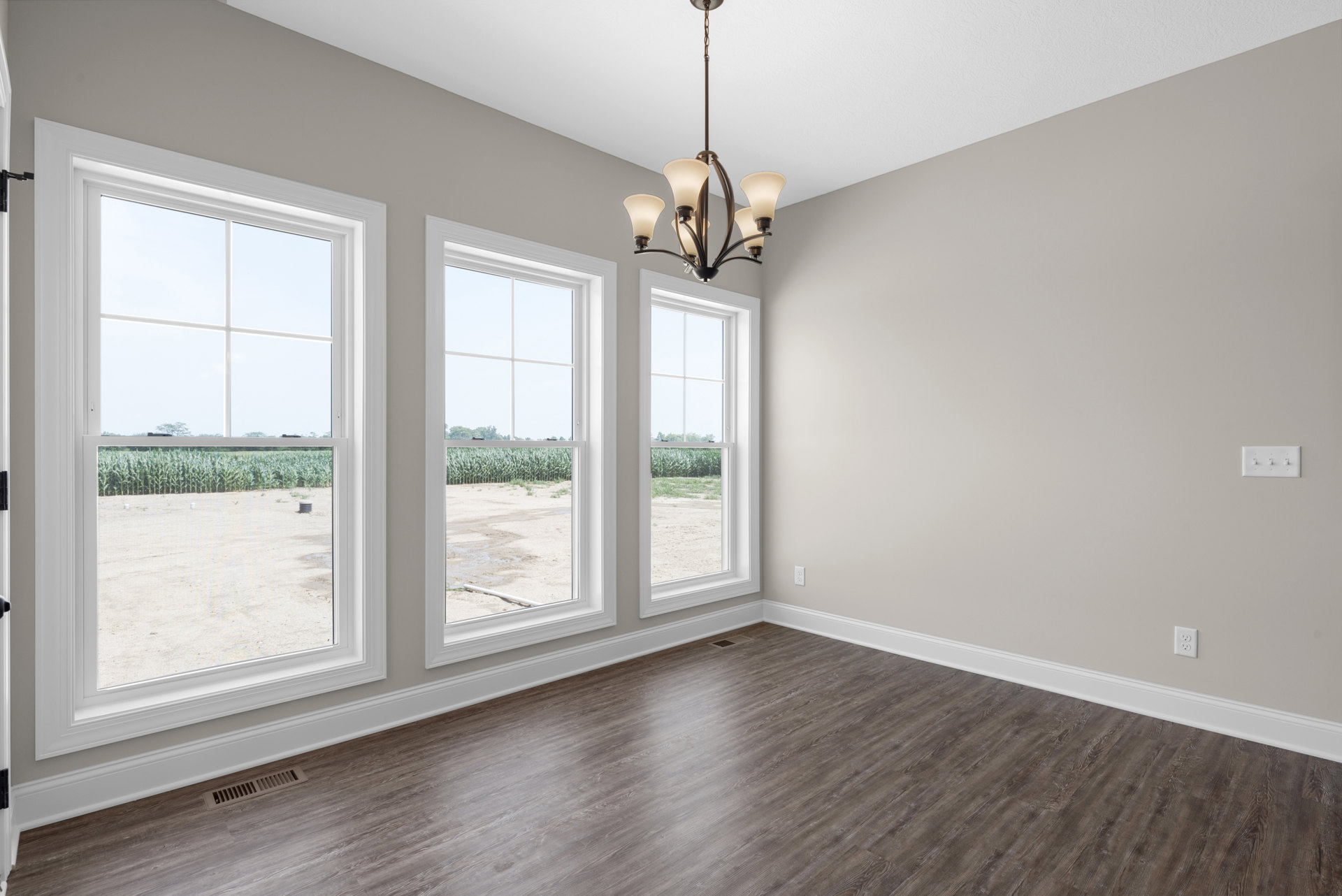Bright room with wood flooring, large windows overlooking a cornfield, white-shaded chandelier, wall vent, and crown molding.