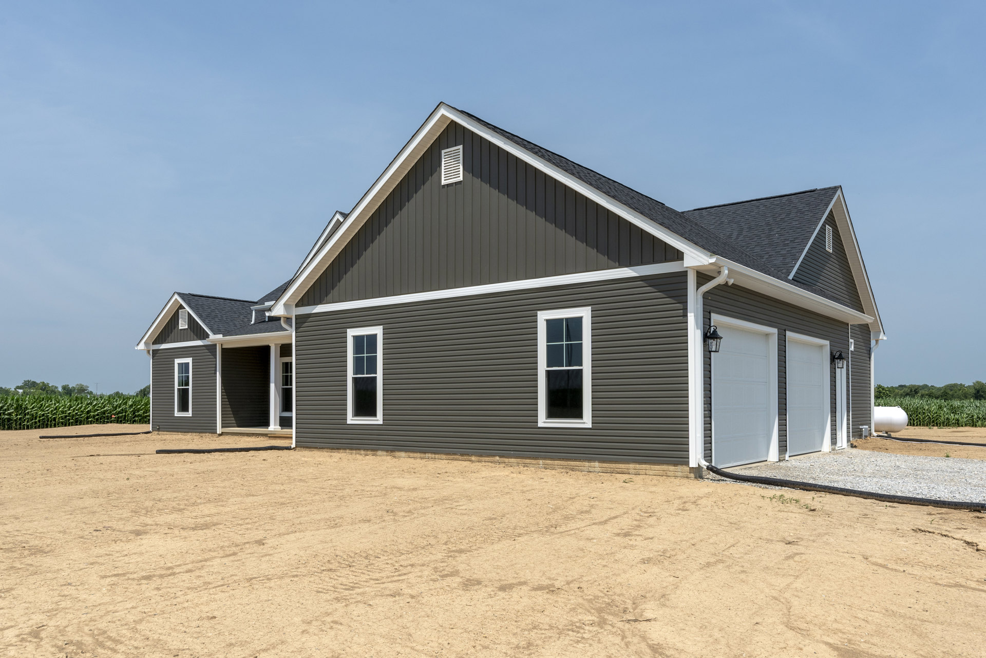 Two-story home with attached garage, white-framed windows, gray siding, shingled roof, dirt driveway, and open sky in background
