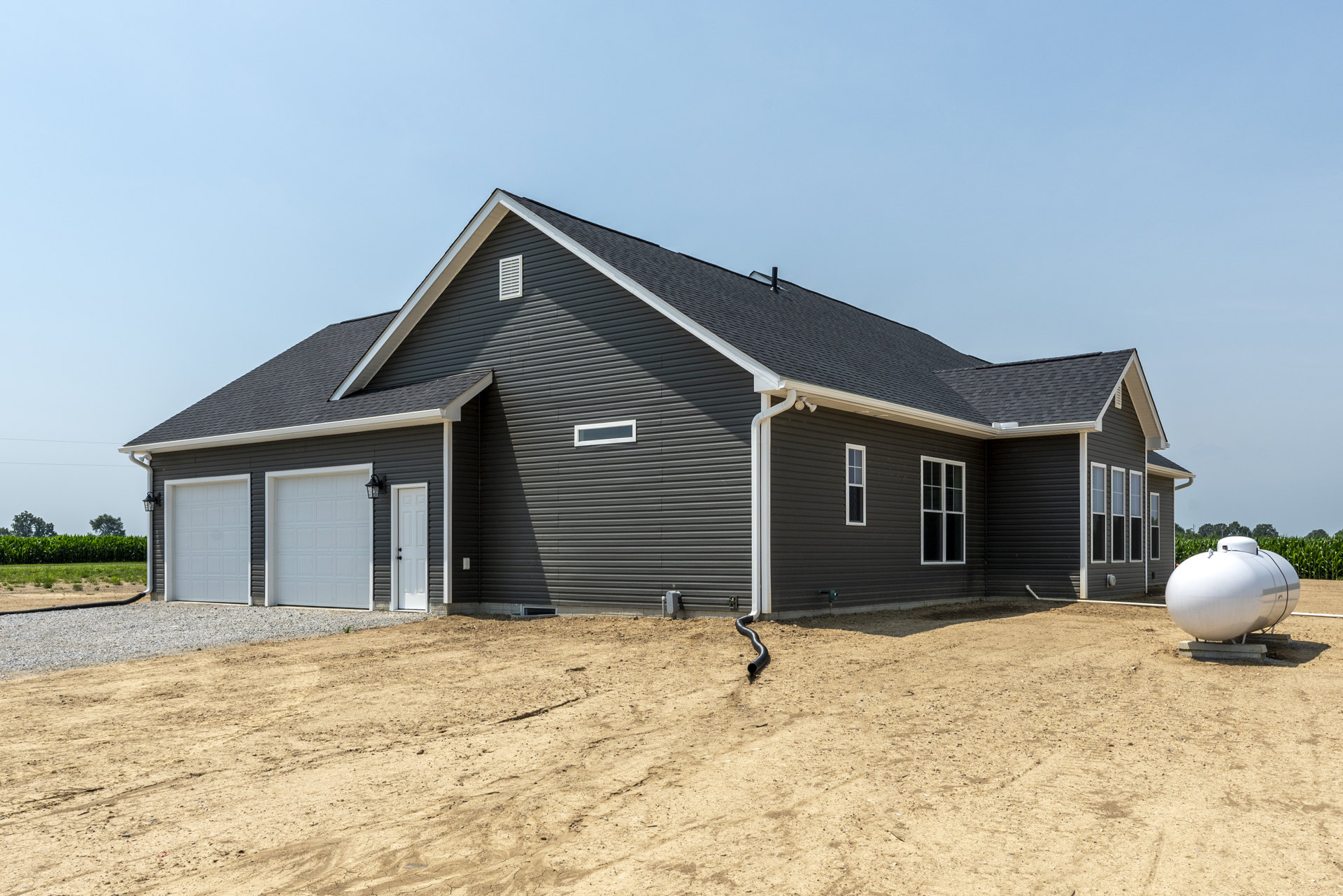 Single-story home with white garage door, black accent wall, large white water tank on metal stand, dirt yard with scattered black piping, sandy ground, and clear sky.