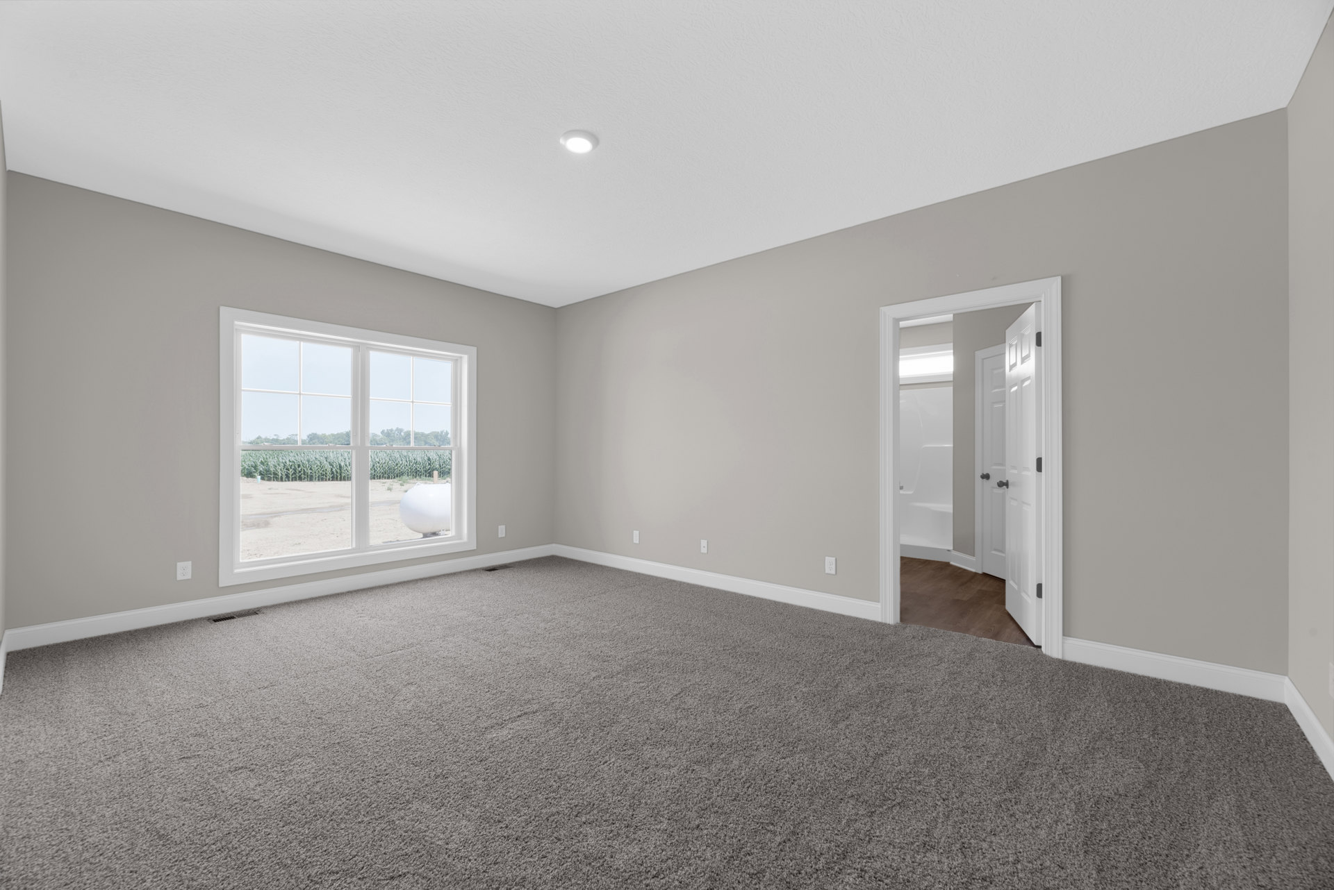 Open door leading to a carpeted room with a window overlooking a cornfield, white round object on the floor, adjacent bathroom featuring white doors and a bathtub, laminate and