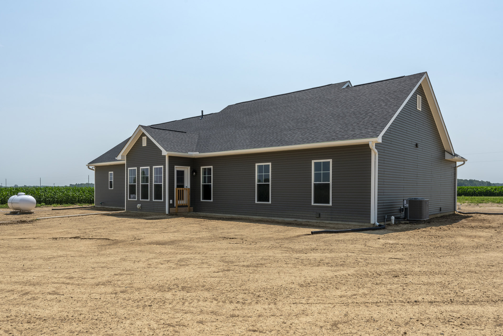 Single-story house with light-colored siding, white-framed window, large air conditioner unit, white water tank on metal stand, dirt yard with scattered objects, clear sky above