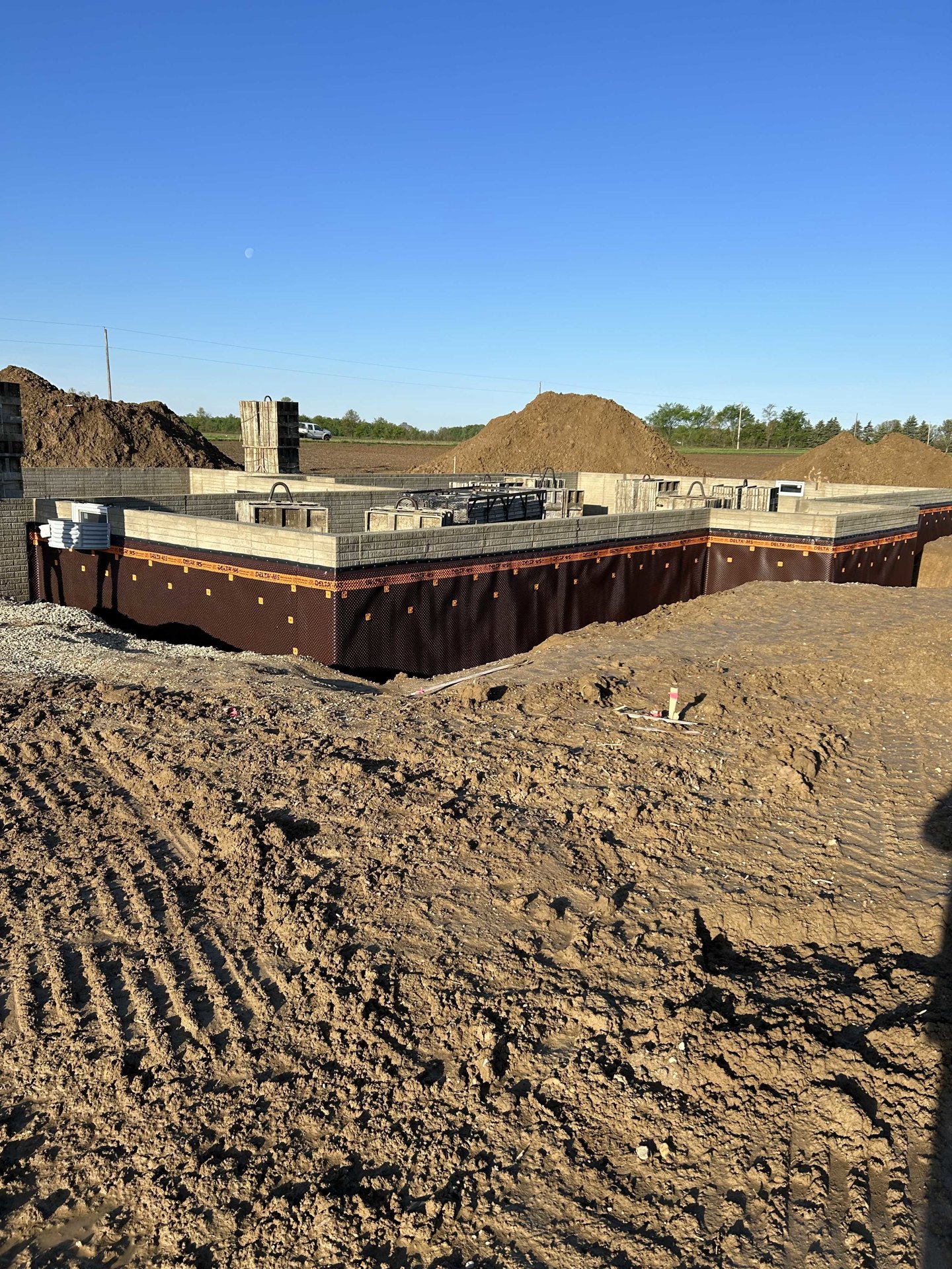 Partially built house with exposed framing, surrounded by dirt piles and construction materials, wooden crate secured with rope, temporary fencing along perimeter, open sky above