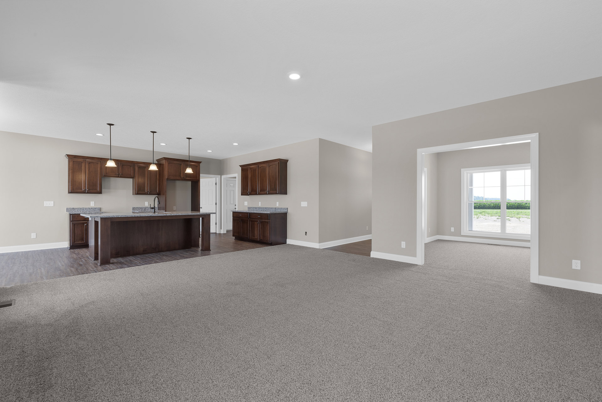 Spacious open-plan room featuring a modern kitchen with wood cabinetry, laminate flooring, a countertop with sink, carpeted dining area, and large window overlooking a grassy