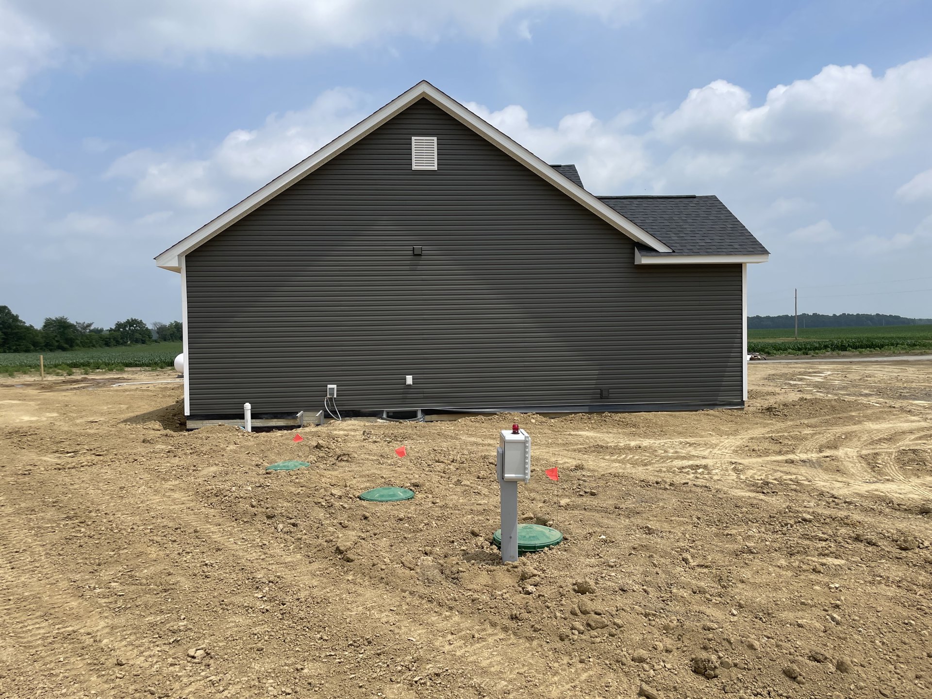 Single-story rural home with light-colored siding, metal roof, and green utility box set on bare dirt lot under partly cloudy sky.