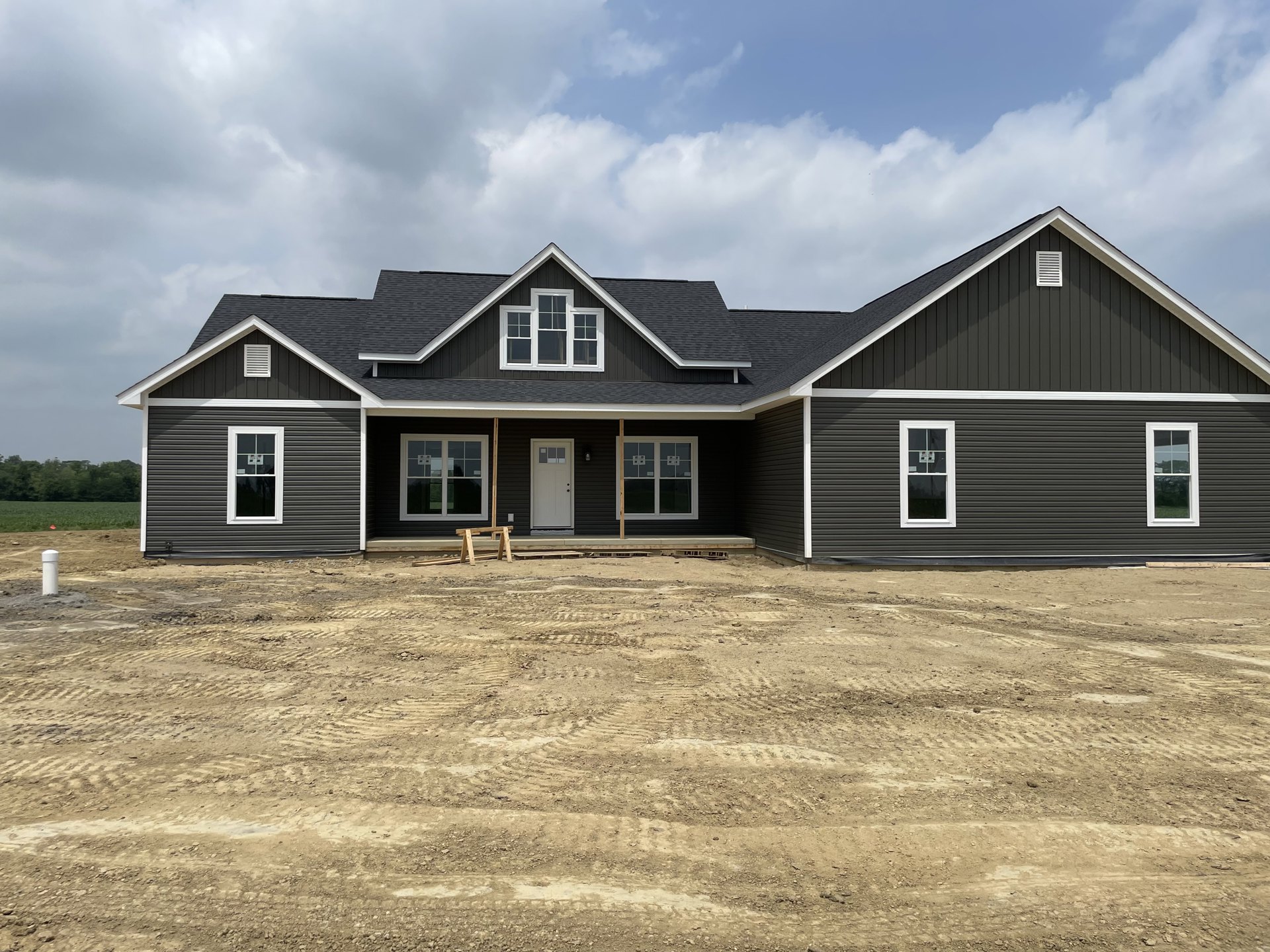 Two-story house under construction with white siding, covered porch, white framed windows, and dirt lot in foreground under cloudy sky