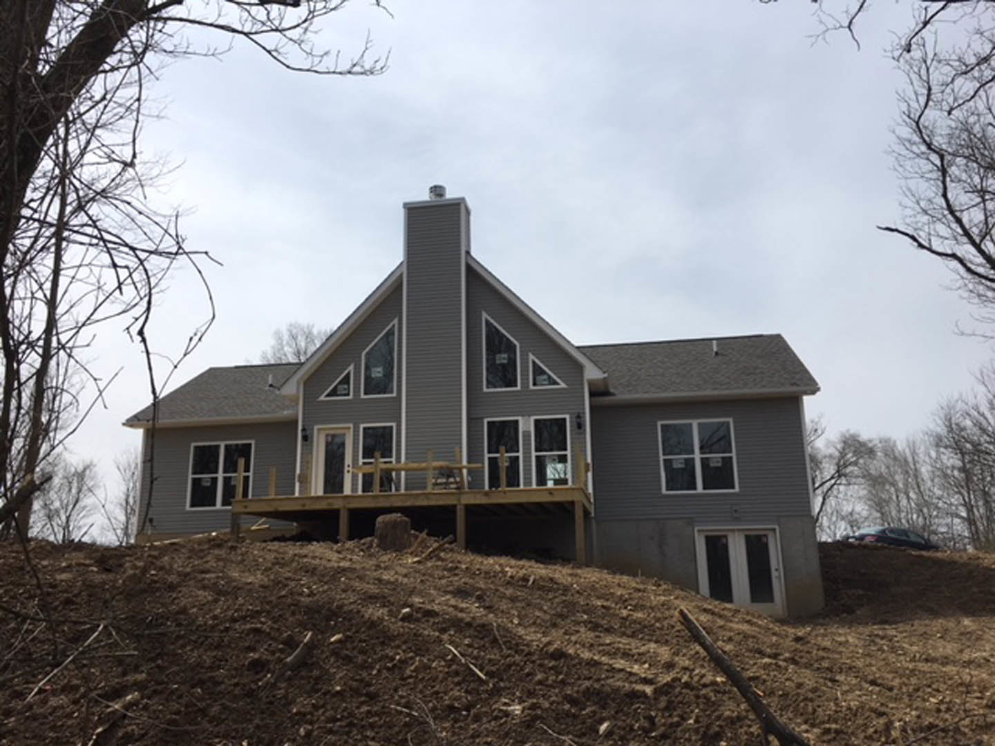 Partially built house with exposed wooden framing and deck, situated on a dirt hill, white-framed window, construction sign on pole, scattered logs, cloudy sky and trees in