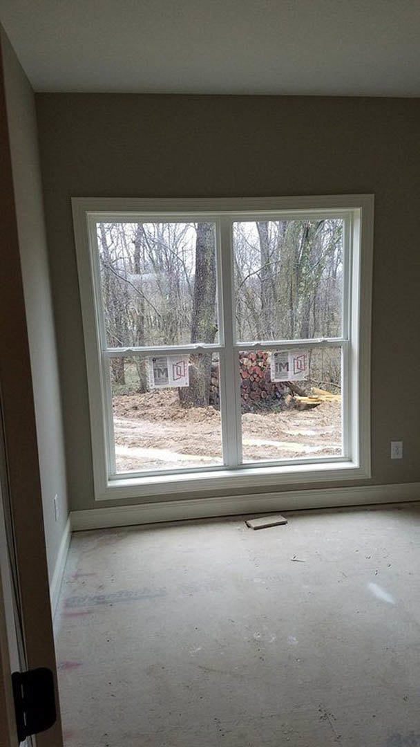 Large window framed by plaster walls, overlooking trees and dirt outside; concrete floor with a single tile, stack of logs near the window, and a close-up of paper on the floor.