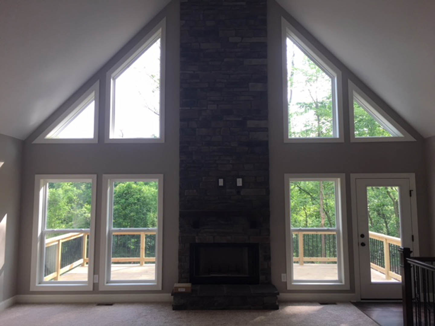 Modern living room with black-framed fireplace set in a brick accent wall, large windows with white trim, hardwood floors, and views of trees outside.