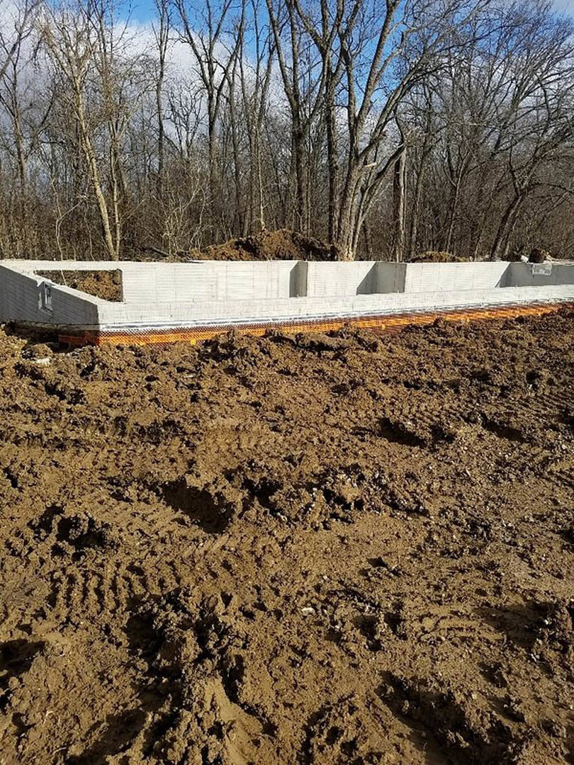 Concrete foundation slab set in a dirt field with tire tracks, bordered by scattered trees under a clear sky