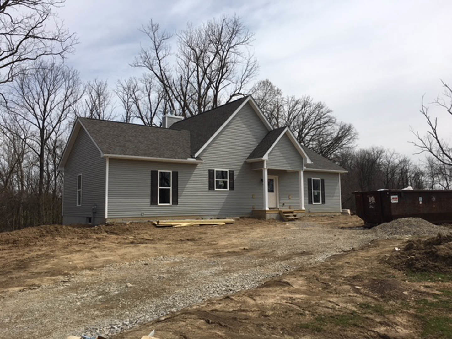 White-roofed house under construction in wooded area, brown dumpster and dirt pile in foreground, white-framed window visible on unfinished siding