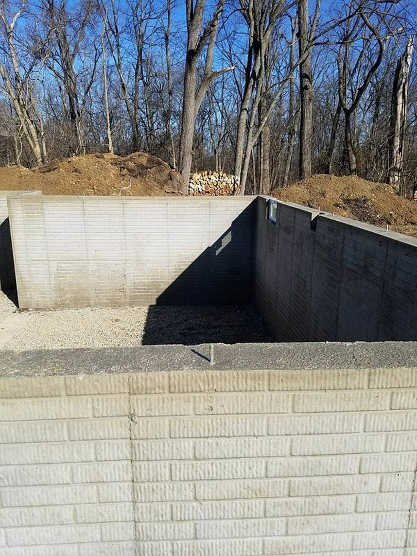 Concrete wall with circular ground opening, white brick accent, blue object resting on wall, building shadow cast across surface, pile of wood nearby, black soil foreground, trees