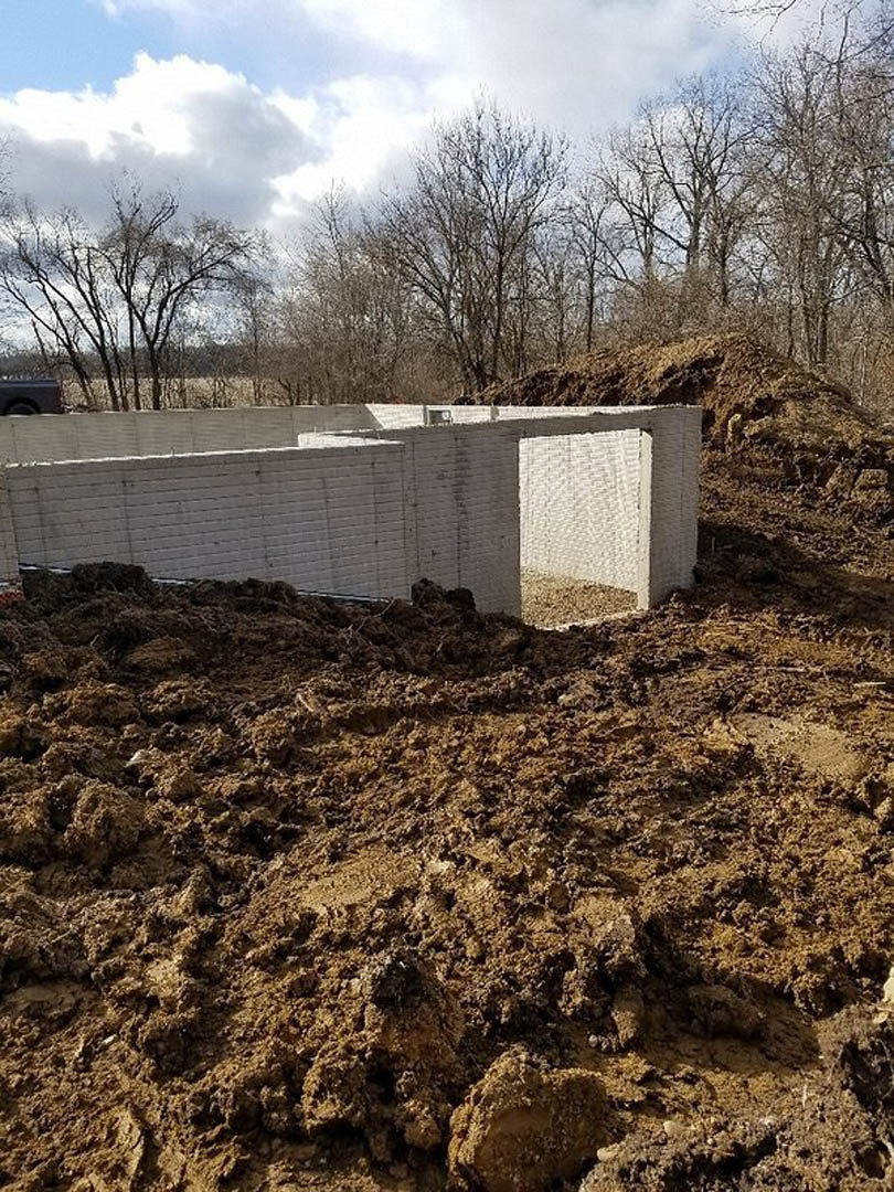 White brick exterior wall with open window framing, piles of dirt in foreground, surrounding trees and cloudy sky in background