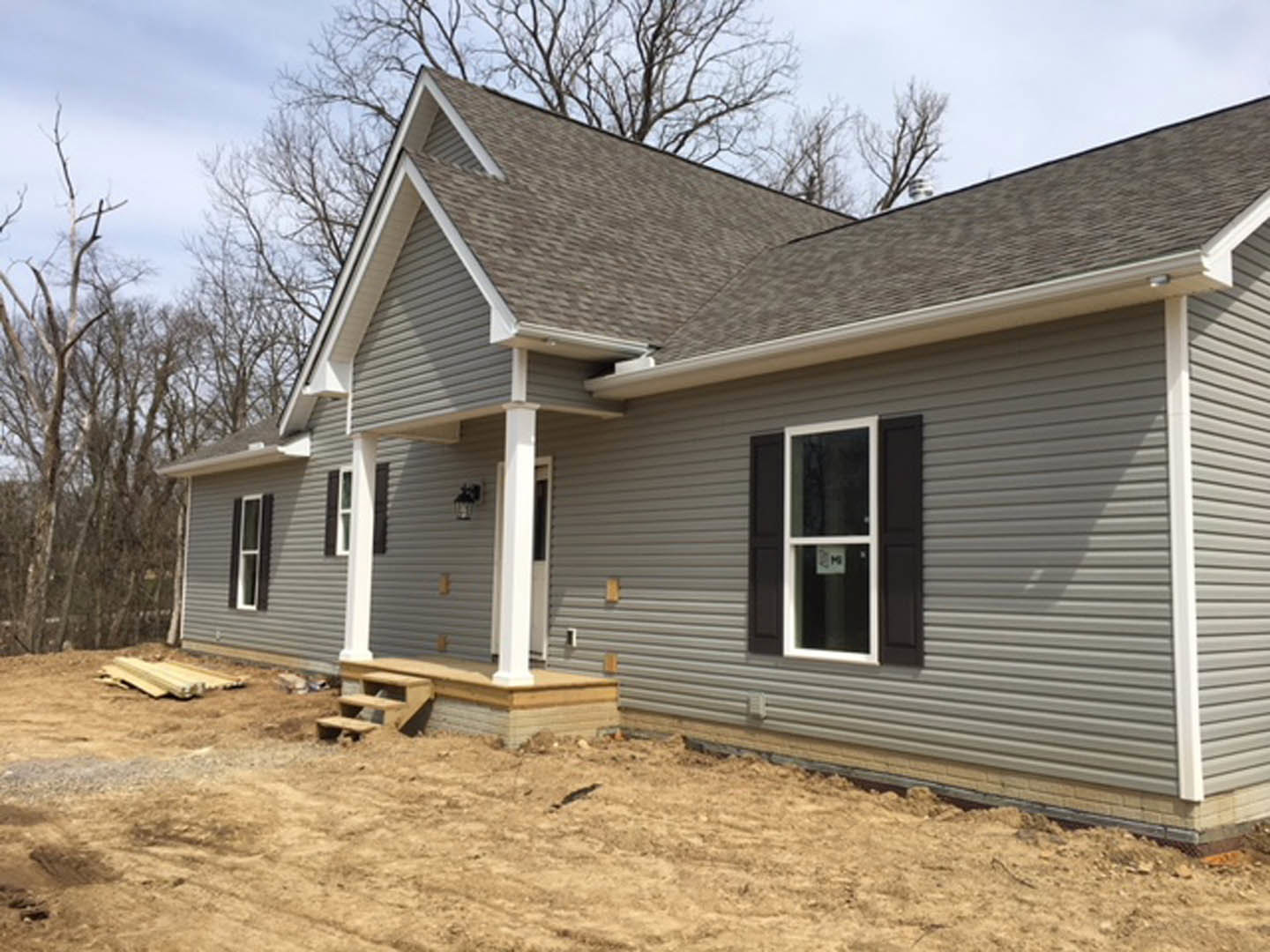 Two-story house under construction with light siding, covered front porch, dirt yard, and mature trees in background