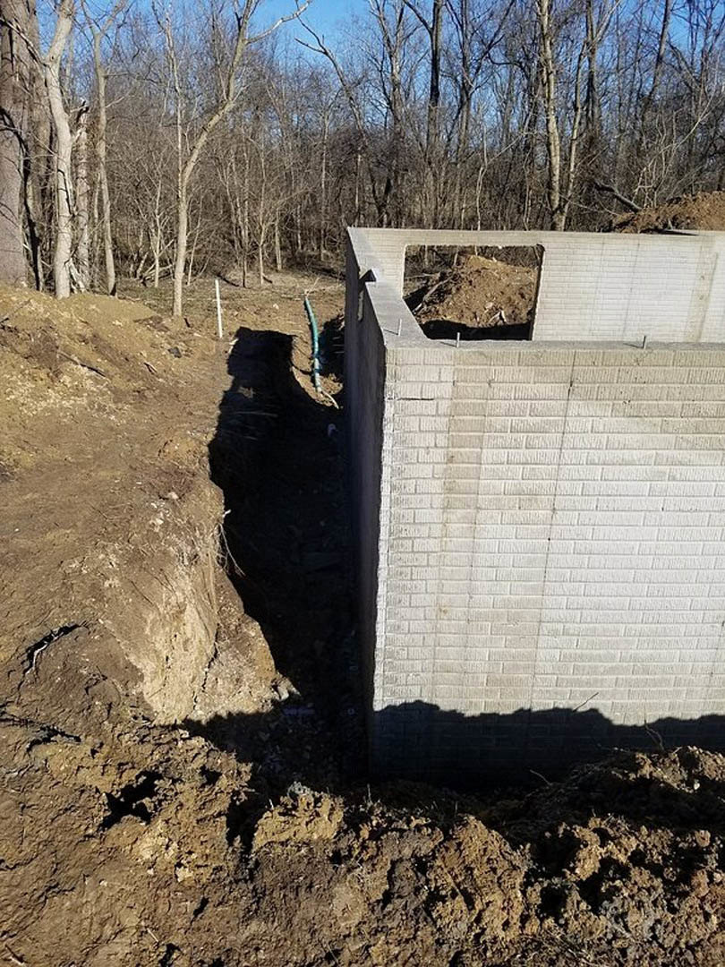 Concrete retaining wall with circular opening, surrounded by soil and dirt, outdoor setting with trees and sky in background