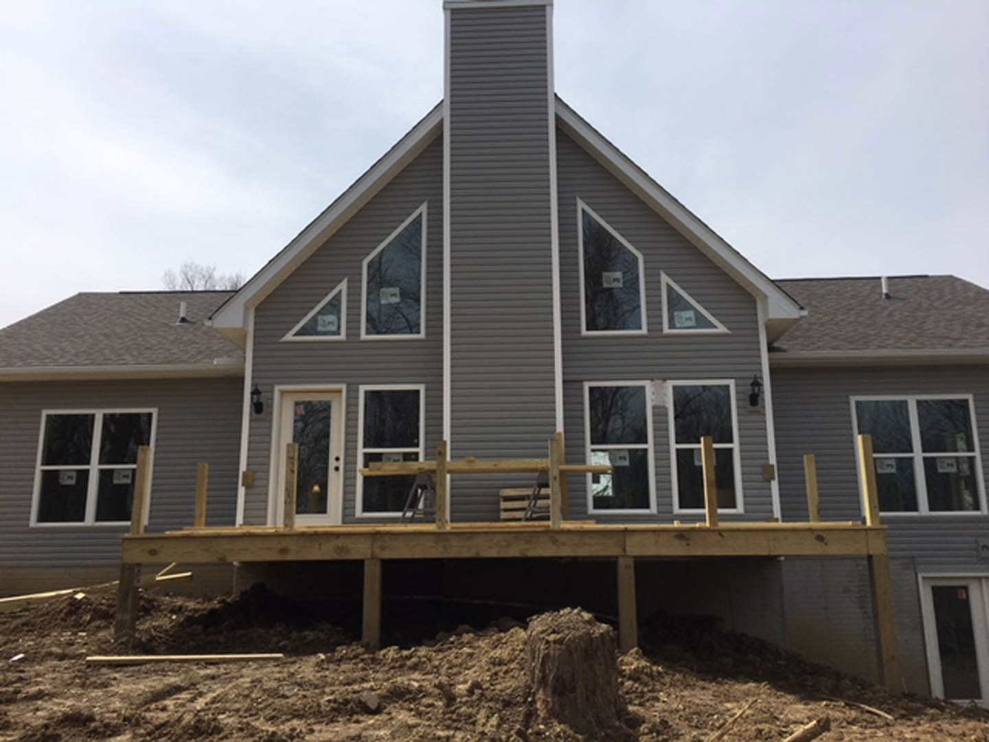 Partially built house with exposed wooden deck, pile of dirt in foreground, white framed window, light siding, and visible roof structure under clear sky