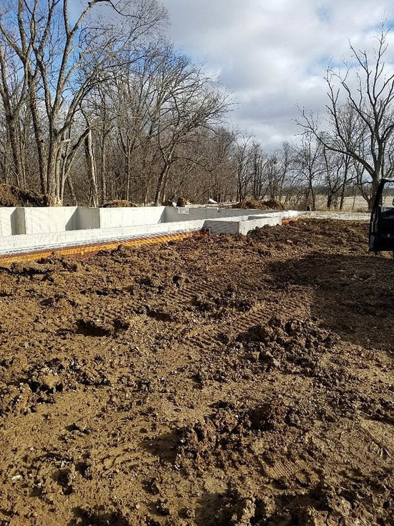 Dirt field bordered by leafless trees and a concrete wall under a cloudy sky