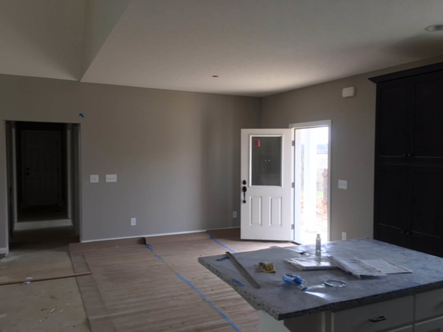 Modern kitchen featuring light tile flooring, white cabinetry, black countertop, stainless steel sink, and a white door with glass window; table holds a face mask and bottle of