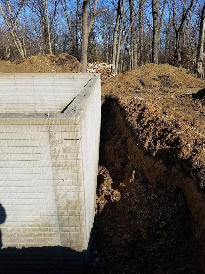 Concrete block positioned beside a freshly dug hole in dirt, surrounded by soil and outdoor landscape with trees visible in the background.