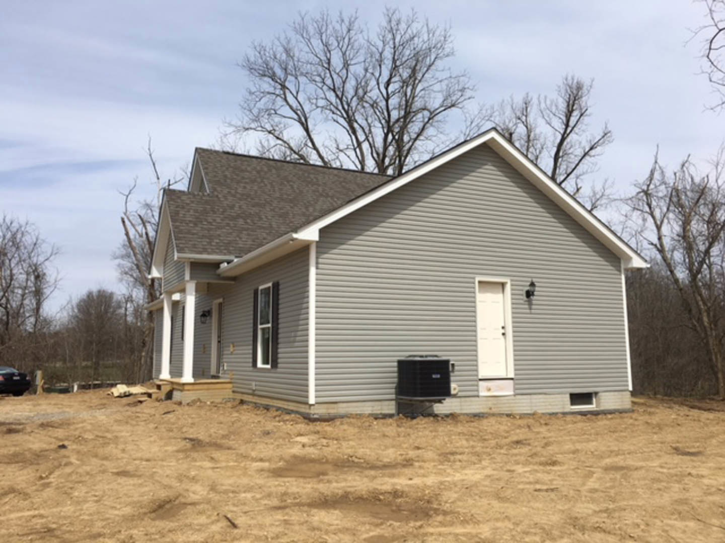 Partially built house with exposed framing, white door, black utility box, dirt yard, and trees in the background