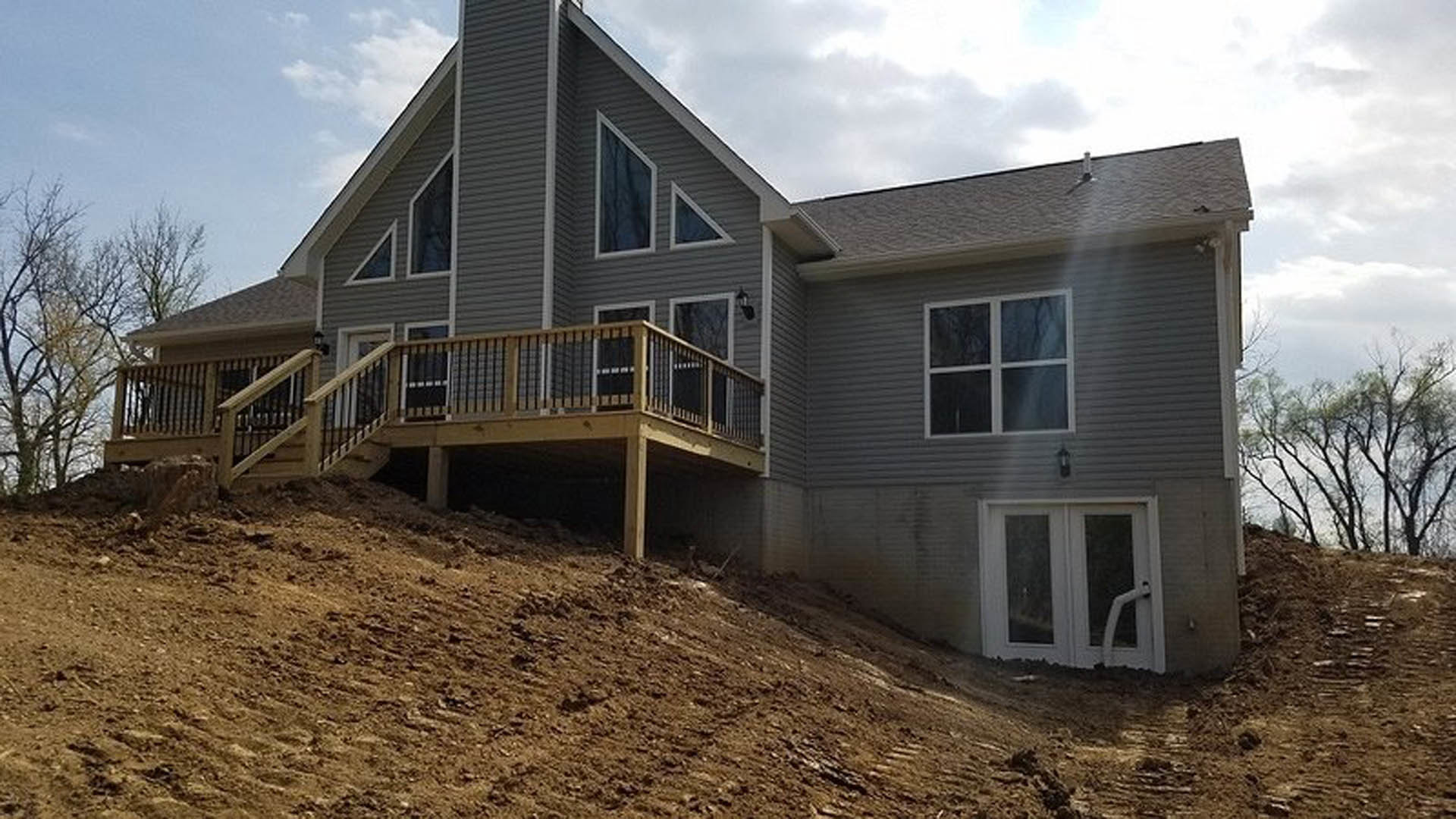 Two-story house with light siding, wooden deck and stairs, white framed windows, glass-paneled door, attached garage, triangular window above entry, surrounded by trees under
