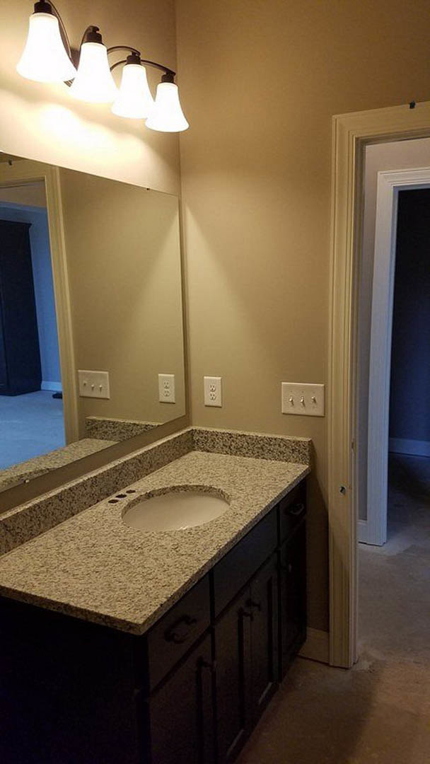 Granite bathroom countertop with white vessel sink featuring black rim, wall-mounted row of lights above, white cabinetry below, chrome faucet and accessories visible