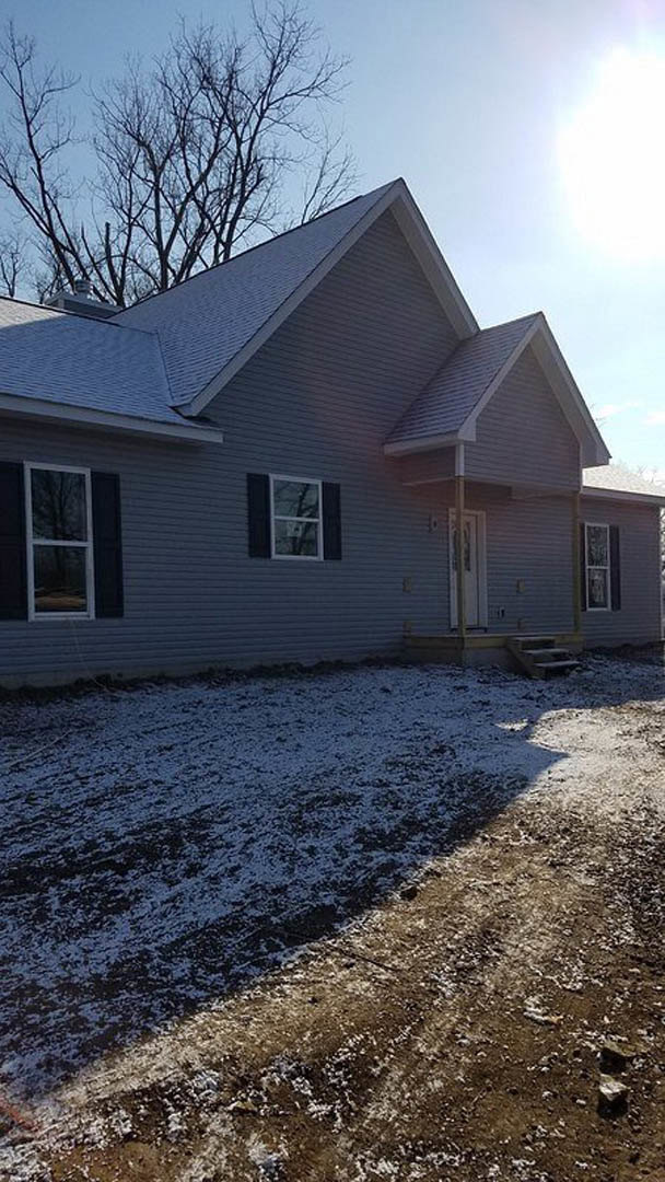 Two-story house with white siding and large windows, snow covering the ground and roof, bare tree branches near the building, blue sky in the background