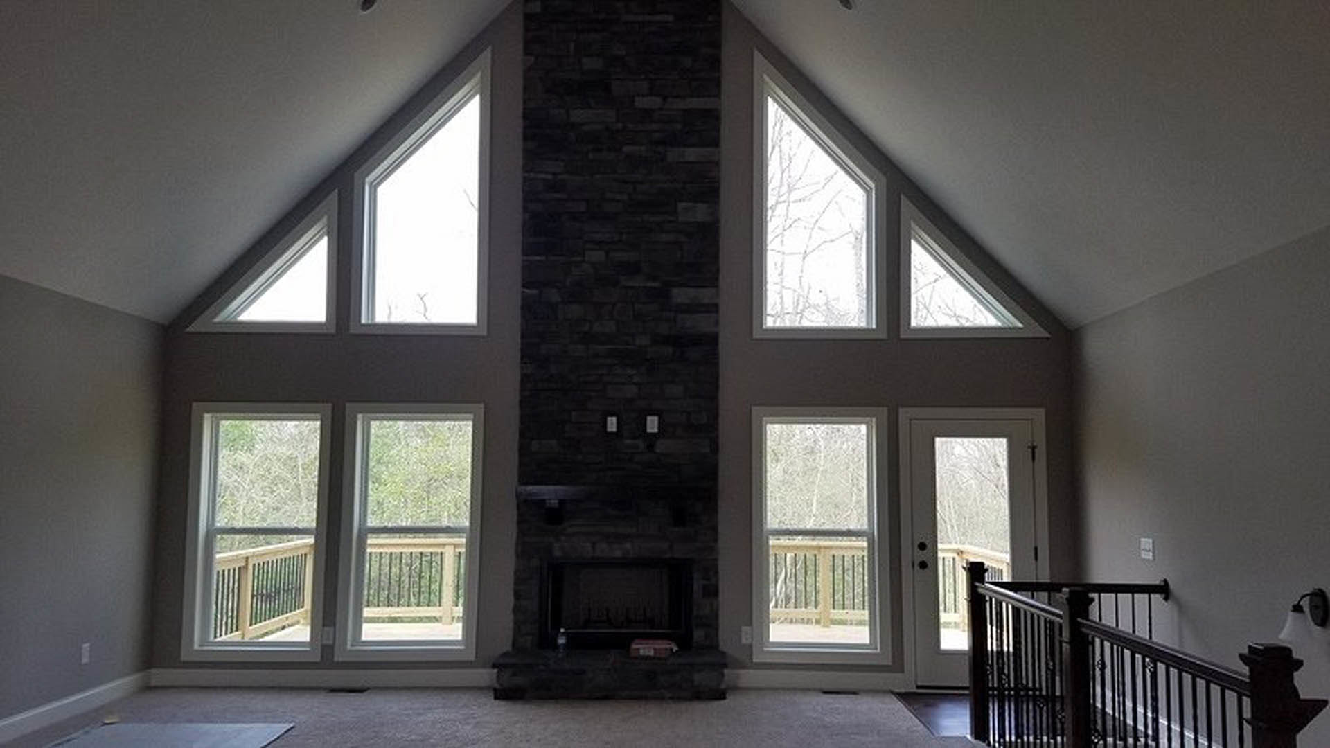 Spacious living area featuring a stone fireplace, hardwood floors, prominent staircase with wooden railing, large windows allowing natural light, white walls with crown molding