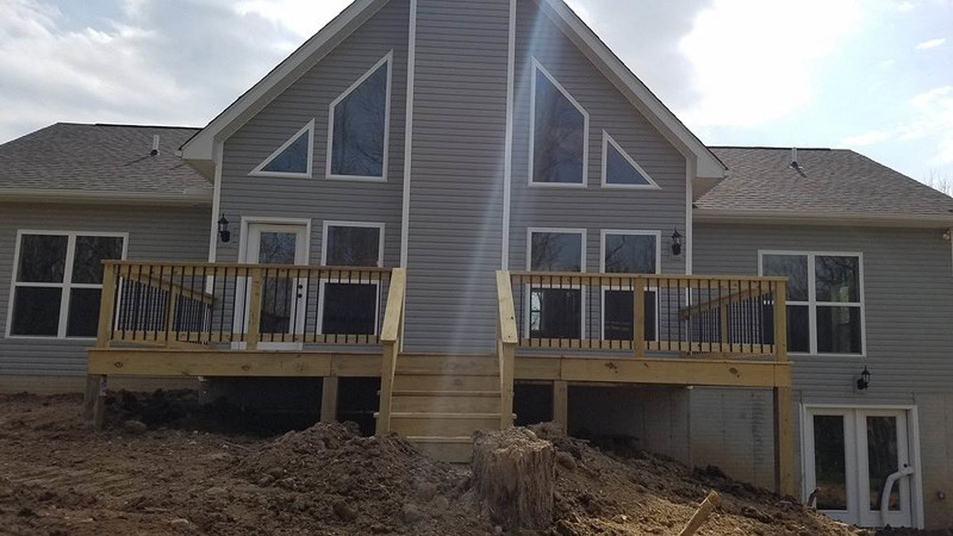 Two-story home with light siding, elevated wooden deck and staircase, metal railing, sash windows, and a large pile of dirt beside the stairs.