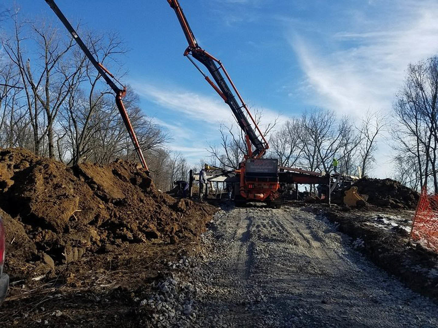 Modern home construction site with crane, dirt piles, rocks, and surrounding trees under cloudy sky