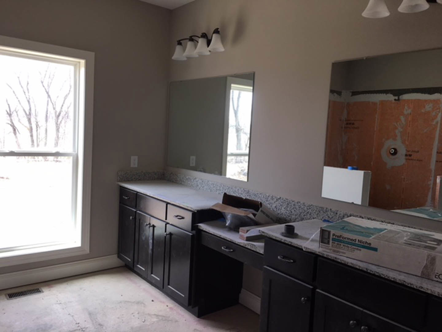 Bathroom with white countertop, rectangular mirror above sink, light fixture with white shade, window showing trees outside, white walls, cabinetry and drawers below counter, vent