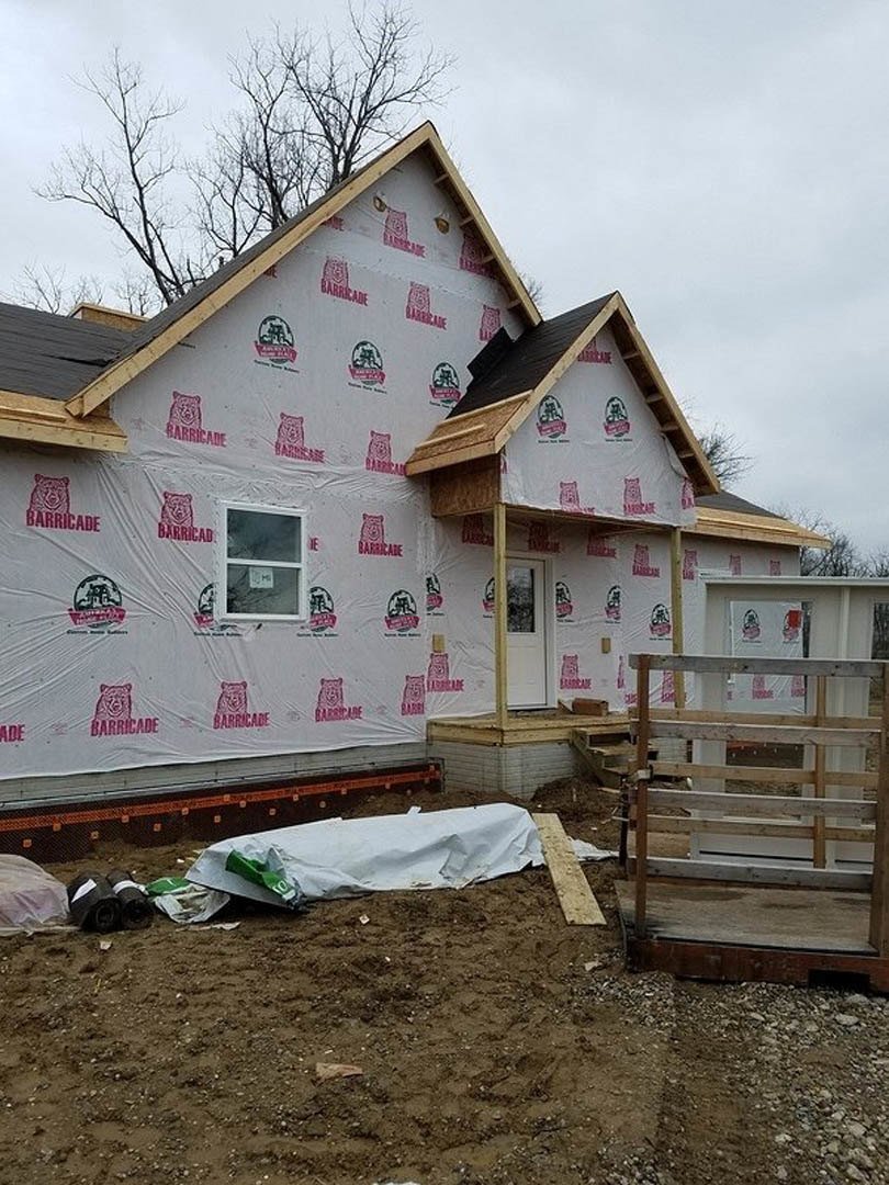 Wood-framed house under construction with plastic sheeting, white door and window, wooden fence enclosing site, white paper on ground, cloudy sky overhead.