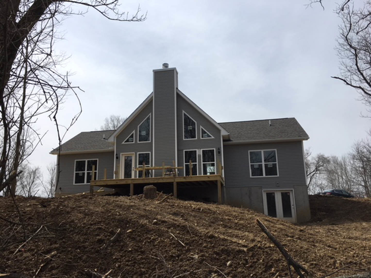 Partially built house on a dirt hill with exposed wooden deck, broken window glass, and white double doors with glass panels; scattered logs and construction materials visible