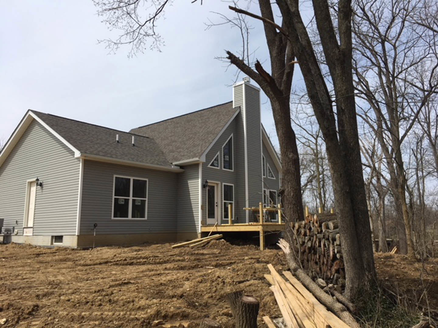 Partially built house with exposed wooden framing, white window frames, unfinished siding, pile of lumber on grassy yard, tree stump and mature tree nearby