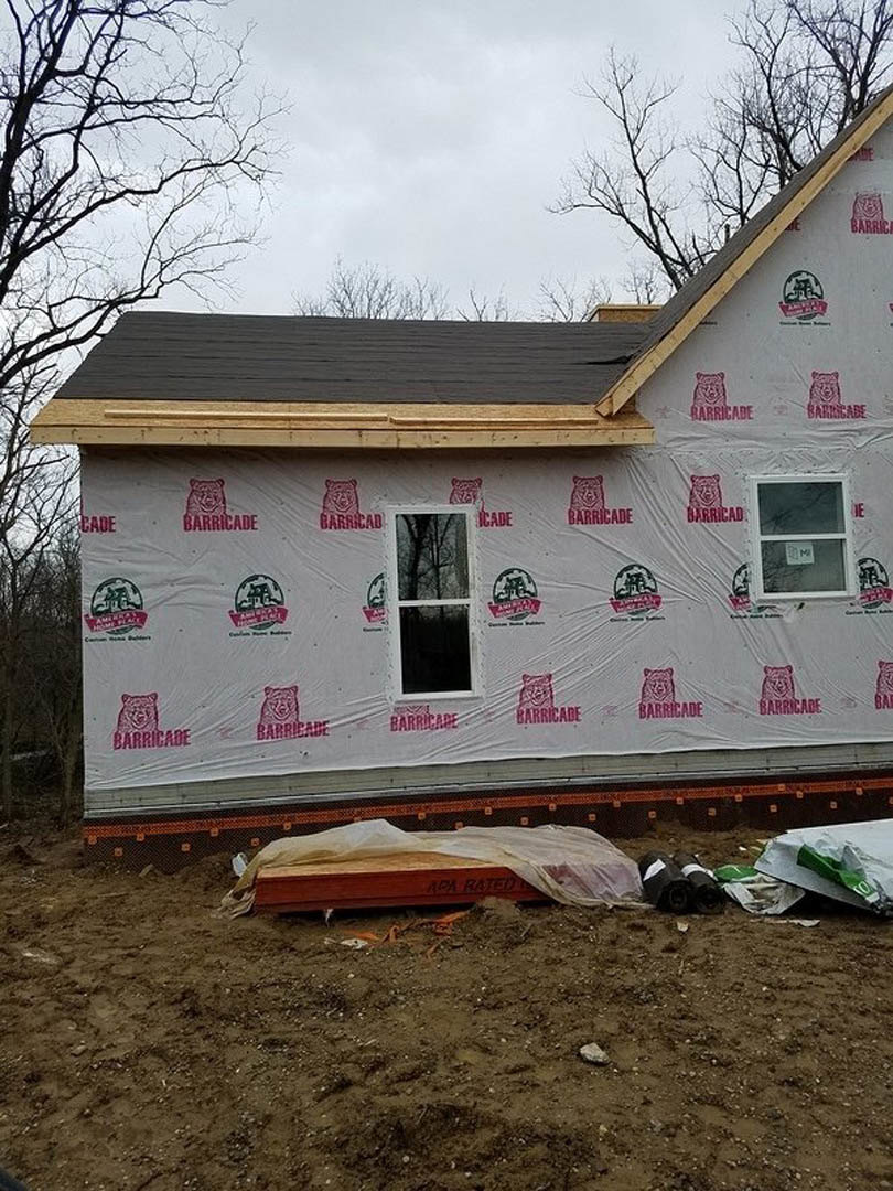 Partially constructed home exterior with plastic sheeting covering windows and siding, white-framed window, brown box with white paper, white sign on window, visible logo, trees