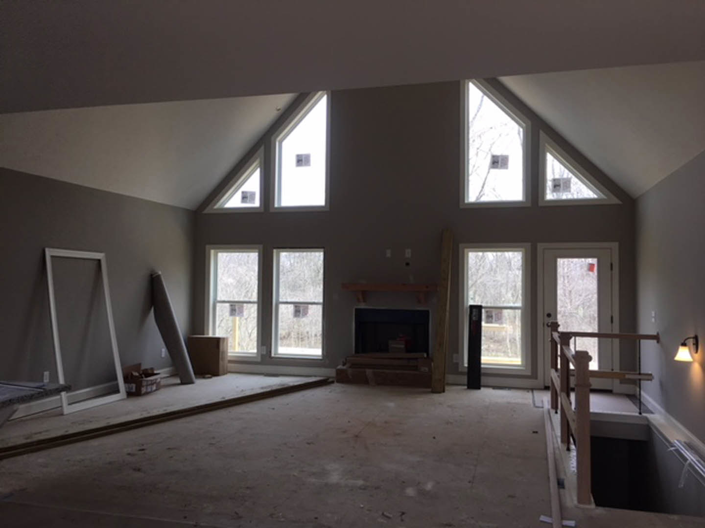 Living room with large windows, white walls, central fireplace with metal surround, wooden railing along hallway, and light-colored flooring.