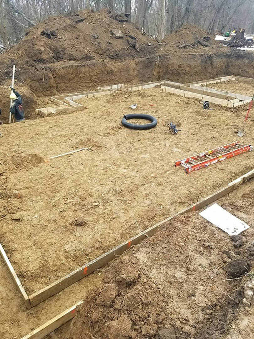 Construction site with exposed soil, scattered ladders, black rubber ring, and blurred figure in jacket near tree and unfinished foundation