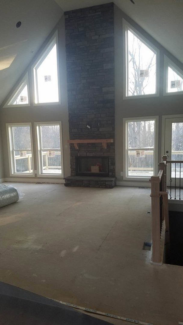 Living room with red brick fireplace, wood mantel, large windows, light-colored walls, and hardwood flooring