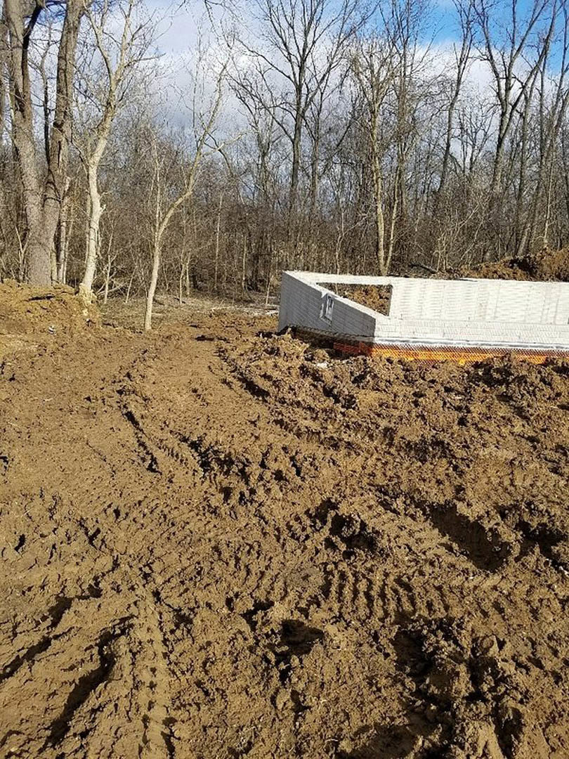 White shipping container set on muddy dirt field with visible tire tracks, bordered by metal fence and surrounded by scattered trees under open sky.