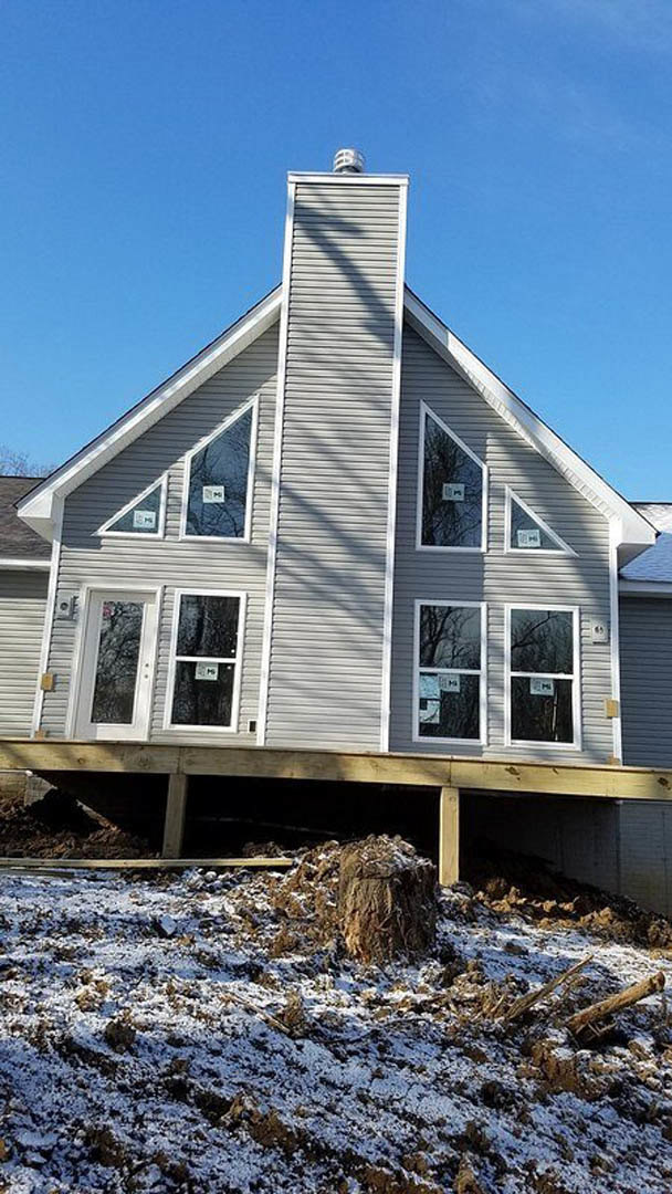 Partially built house with exposed framing, chimney, and siding, surrounded by snow; window displays a white sign, stump and sign on pole visible in foreground