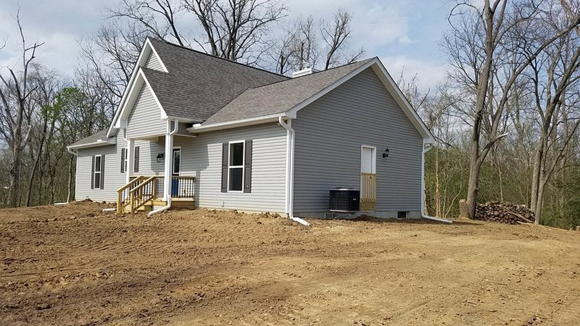 Two-story house with light siding, large windows, and a dirt yard surrounded by mature trees under a clear sky