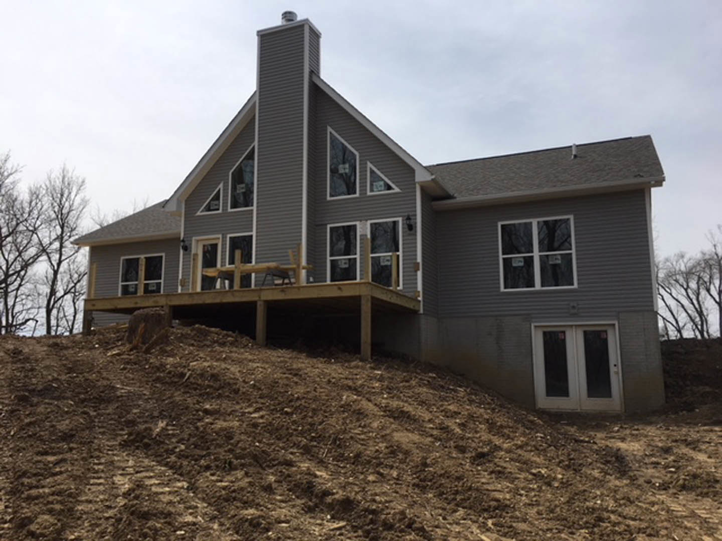 Wood-framed house under construction with glass double doors, white-framed window, elevated wooden deck, and dirt hill in foreground
