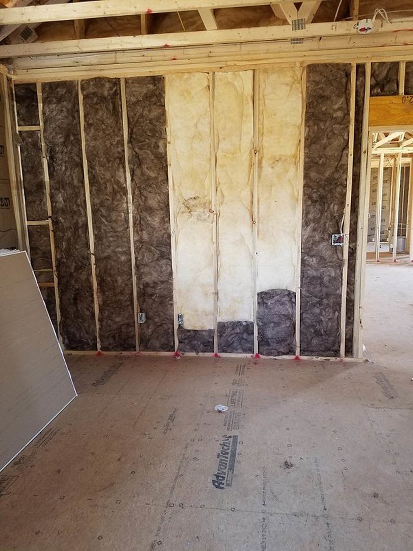 Framed wall with exposed fiberglass insulation, wood beams, and white composite sheathing; unfinished floor and partial view of door with manufacturer logo.