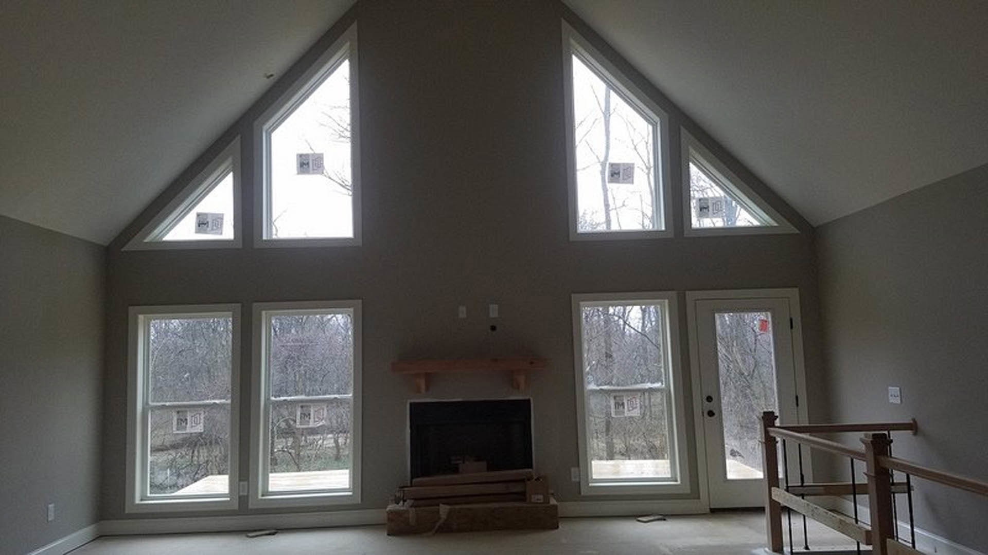 Living room with stone fireplace, large windows, hardwood floors, and white walls; modern staircase with metal railing visible in foreground.